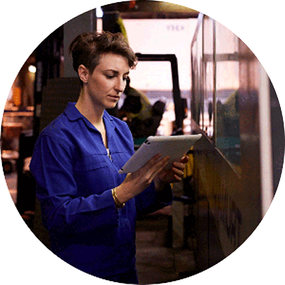 A woman in a blue jumpsuit holds a tablet while standing next to a wall in a warehouse setting