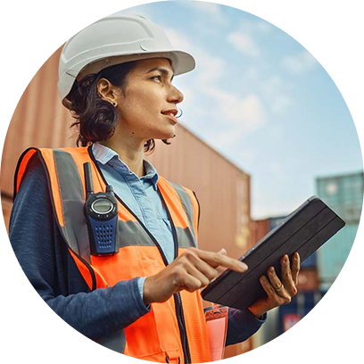 A woman in a hard hat and safety vest holds a tablet while looking up, standing near shipping containers